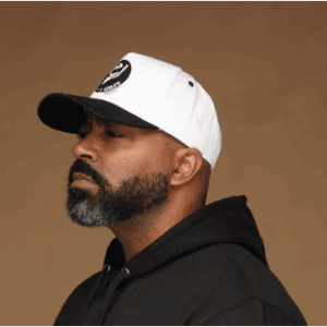 Profile of a bearded man wearing a white cap and black hoodie.